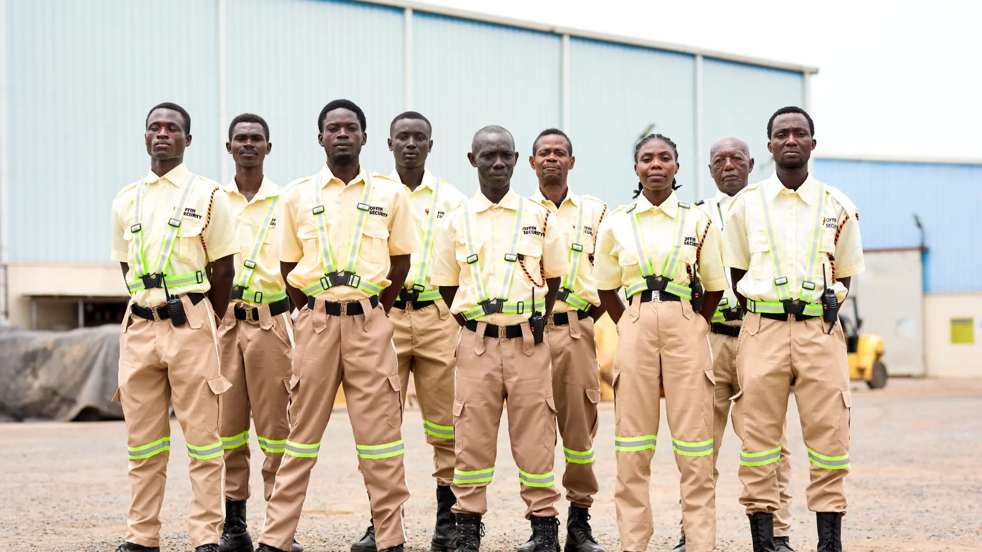 Offin Security guards in cream shirts, khaki trousers and high-visibility harnesses standing at attention in formation at an industrial site