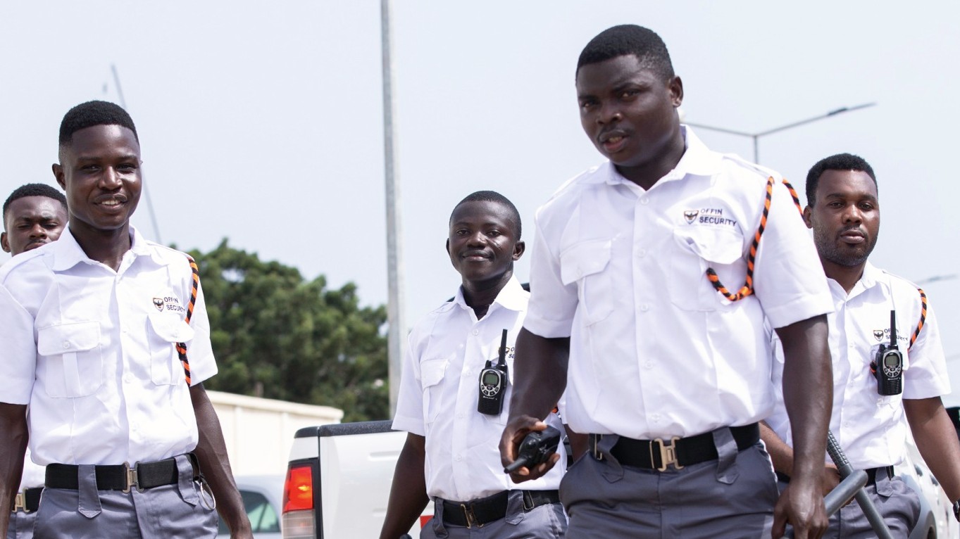 Five Offin Security guards in white shirts and grey cargo trousers walking in formation past a pickup truck, one carrying a radio and a baton
